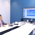 会社の会議室エリアで、常設のスクリーンと、ノートパソコン、iPadを用いて講義を行う粟国氏