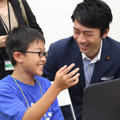 Minecraftコースの中学2年生から、Minecraftについて説明を受ける小泉氏。明瞭な回答と解説に「説明能力が高いね！」と驚く場面も。何かを作る楽しみや今自分が行っていることを熱心に話すようすに、小泉氏は深い感銘を受けていた